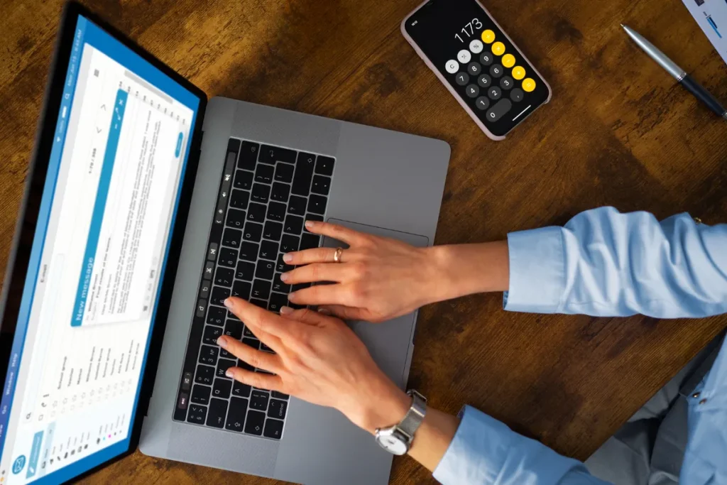Mulher digitando em laptop com calculadora e caneta na mesa, gerenciando saldo remanescente financeiro.