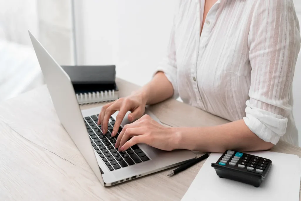 Mulher digitando em um laptop, com calculadora e caderno na mesa, focada em finanças e saldo remanescente.