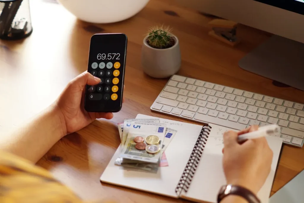 Pessoa calculando finanças em um smartphone, com notas de dinheiro e moedas em um caderno.
