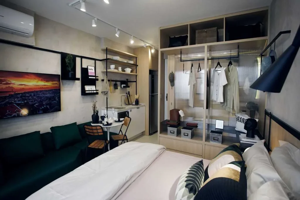 Apartamento studio moderno com cama de casal, sofá verde-escuro, cozinha compacta e guarda-roupa aberto integrado ao ambiente.