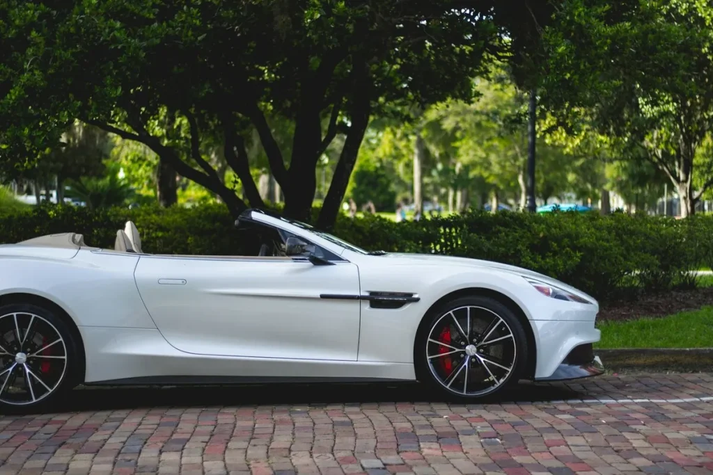 Carro esportivo conversível branco, um Aston Martin Vanquish, estacionado à luz do dia em uma área arborizada.