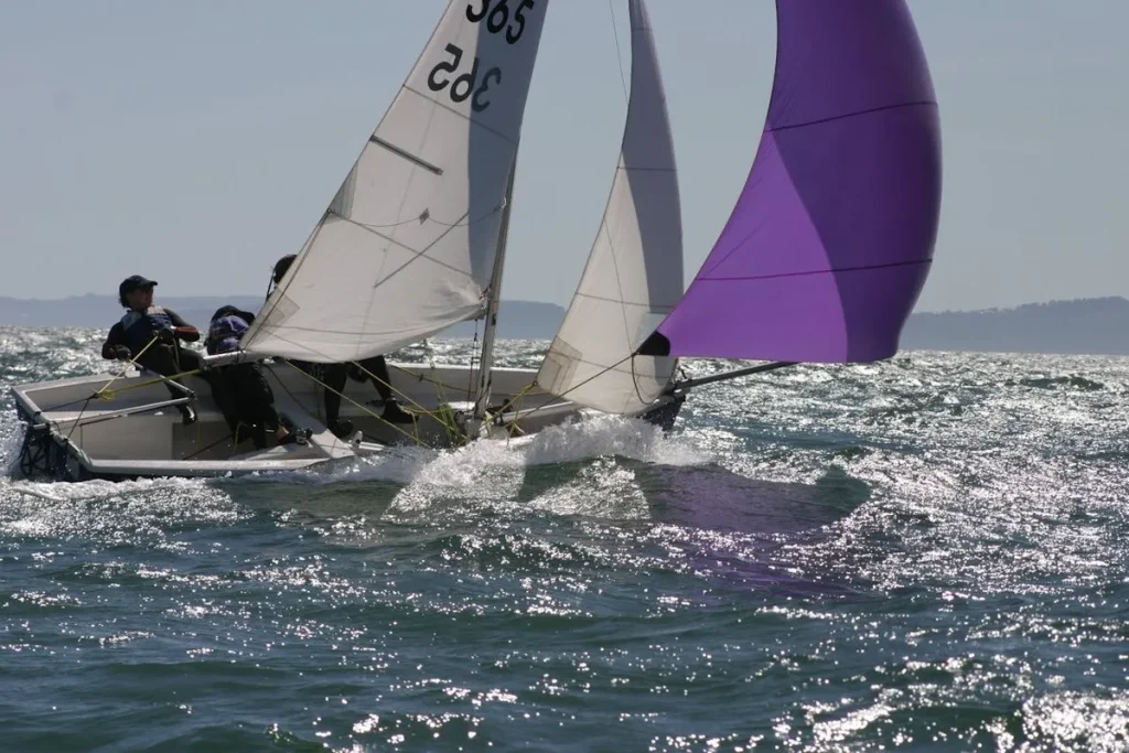 Um pequeno veleiro de regata, com vela principal branca e vela colorida (gennaker) na cor roxa, navegando em águas agitadas com dois tripulantes a bordo. A imagem retrata a velocidade e o dinamismo de uma competição de veleiro.
