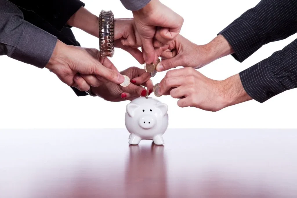Várias mãos de diferentes pessoas depositando moedas em um cofrinho de porco. A imagem simboliza a colaboração financeira e a poupança conjunta, representando a ABAC (Análise de Benefícios e Custos) de investimentos coletivos e ações de poupança em grupo.