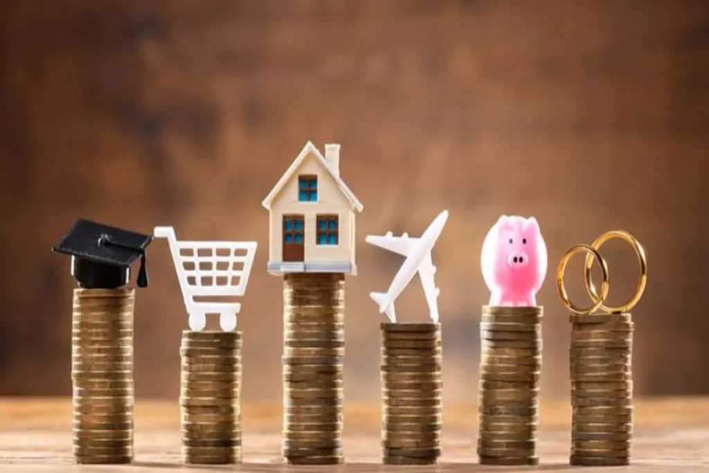 Seis pilhas de moedas de ouro empilhadas em uma superfície de madeira, simbolizando a poupança e o investimento. Cada pilha é encimada por uma miniatura que representa um objetivo financeiro: um capelo de formatura, um carrinho de compras, uma casa, um avião de viagem, um cofrinho de porco e um par de alianças de casamento. A imagem conceitual representa a economia nacional e as prioridades incentivadas pelo Banco Central do Brasil.