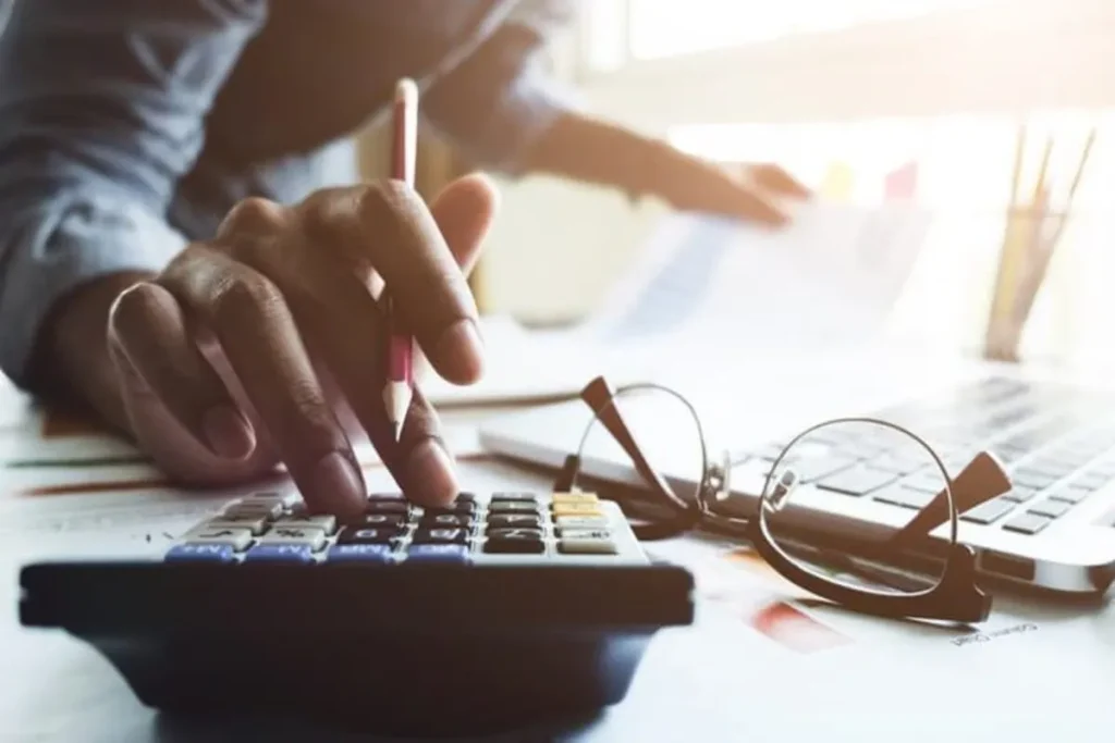 Uma pessoa faz cálculos em uma calculadora, representando a análise de taxas financeiras como o IOF (Imposto sobre Operações Financeiras) para transações de crédito e câmbio.