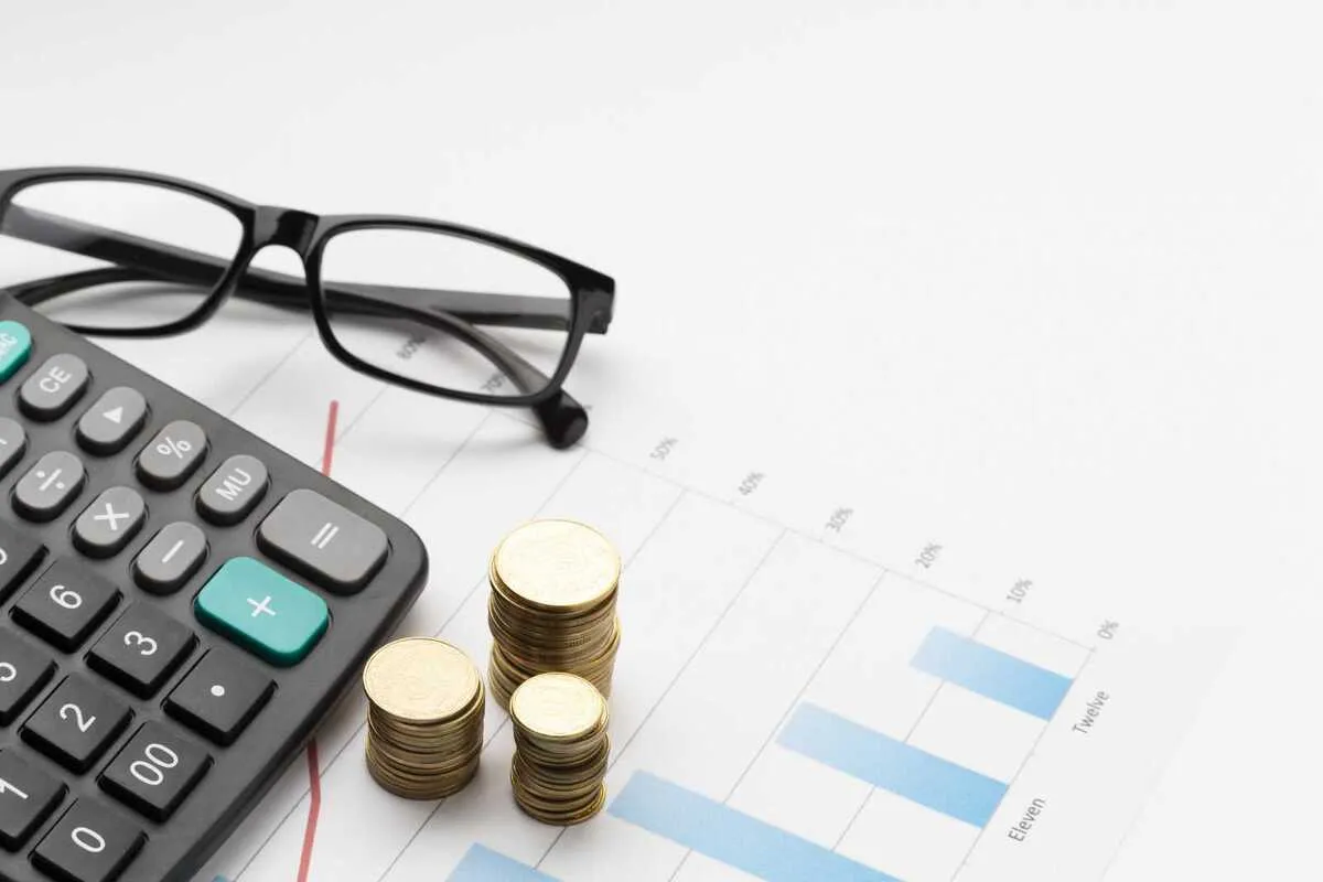 Óculos, calculadora e moedas sobre um gráfico de barras. A imagem representa o cálculo e a análise de investimentos e taxas, como o IOF, que afetam os ganhos financeiros.
