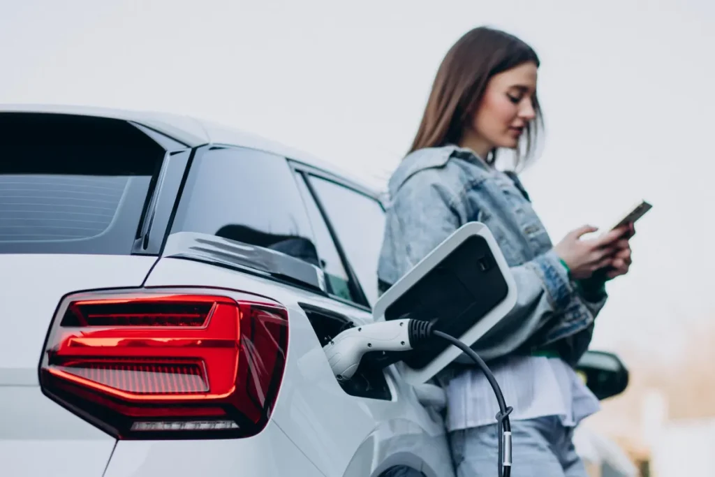 Mulher usando um smartphone enquanto seu carro elétrico está conectado a uma estação de carregamento.