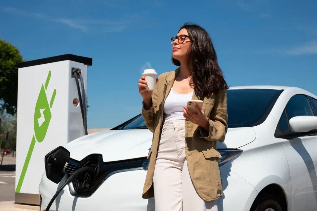 Mulher com óculos de sol e xícara de café, usando o celular ao lado de um carro elétrico em um ponto de recarga.
