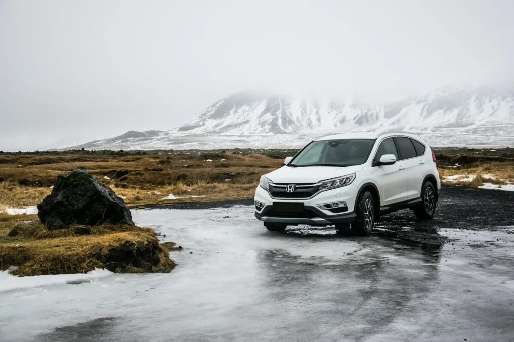 Um SUV branco estacionado em um terreno com gelo e pedras, em frente a uma montanha coberta de neve em um dia nublado.