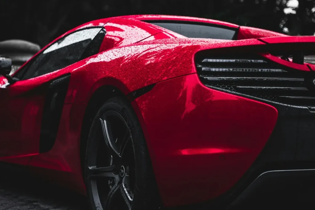 Close-up da traseira de um carro esportivo de luxo vermelho brilhante (possivelmente McLaren) com luzes de freio em LED, molhado pela chuva, simbolizando a aquisição de veículos caros.