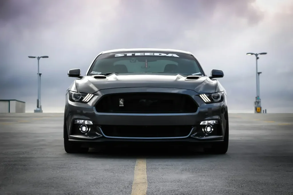 Vista frontal de um Ford Mustang moderno (S550) preto ou cinza escuro, modificado com capô e grade customizados, estacionado em um terraço com postes de luz ao fundo.
