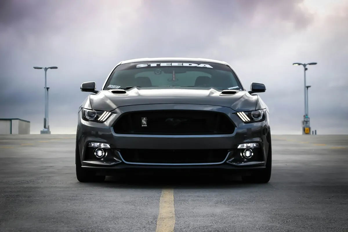 Vista frontal de um Ford Mustang moderno (S550) preto ou cinza escuro, modificado com capô e grade customizados, estacionado em um terraço com postes de luz ao fundo.