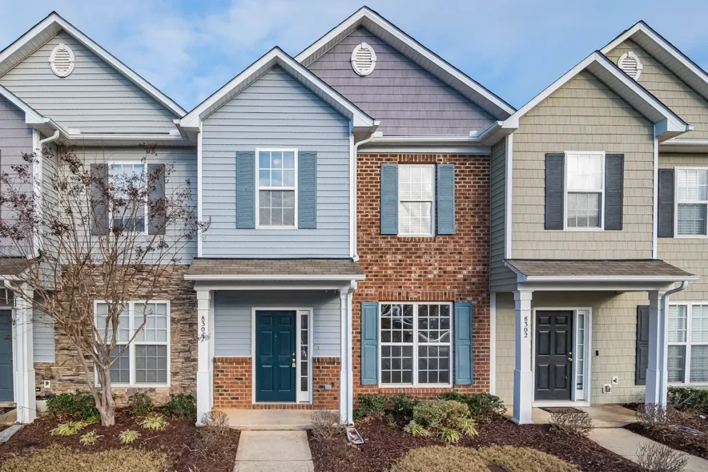 Três casas geminadas em cores pastel (azul, cinza, bege) com fachadas de tijolos e siding, portas escuras e janelas brancas, representando o conceito de Townhouse em um condomínio.