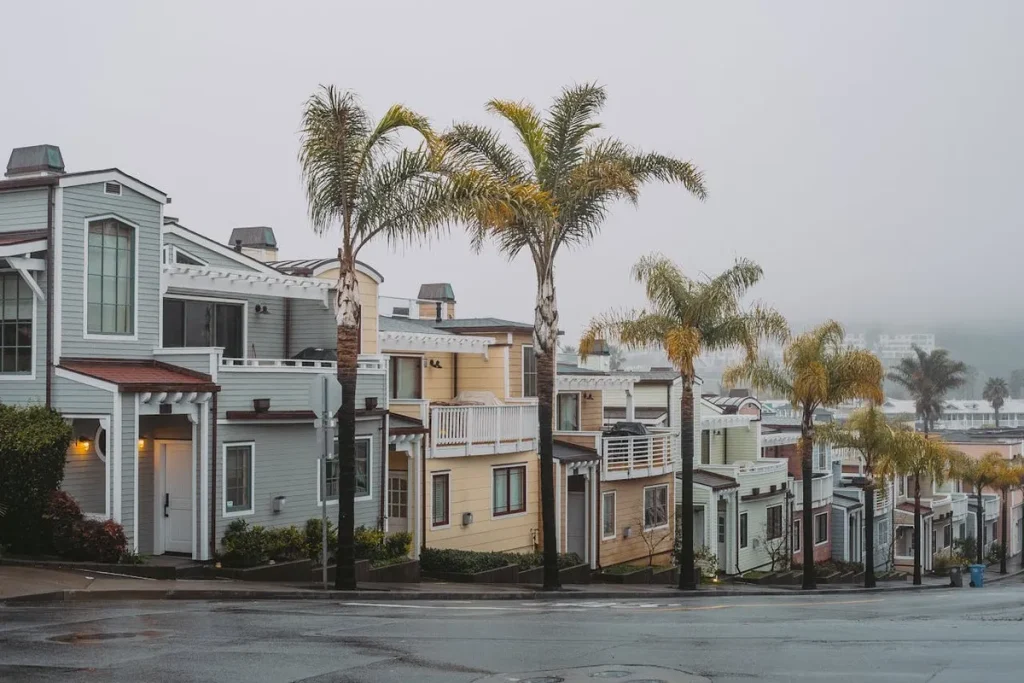 Fileira de casas geminadas (townhouses) em cores pastel na encosta, com varandas e telhados inclinados, separadas por palmeiras, em um dia nublado ou neblina (fog).
