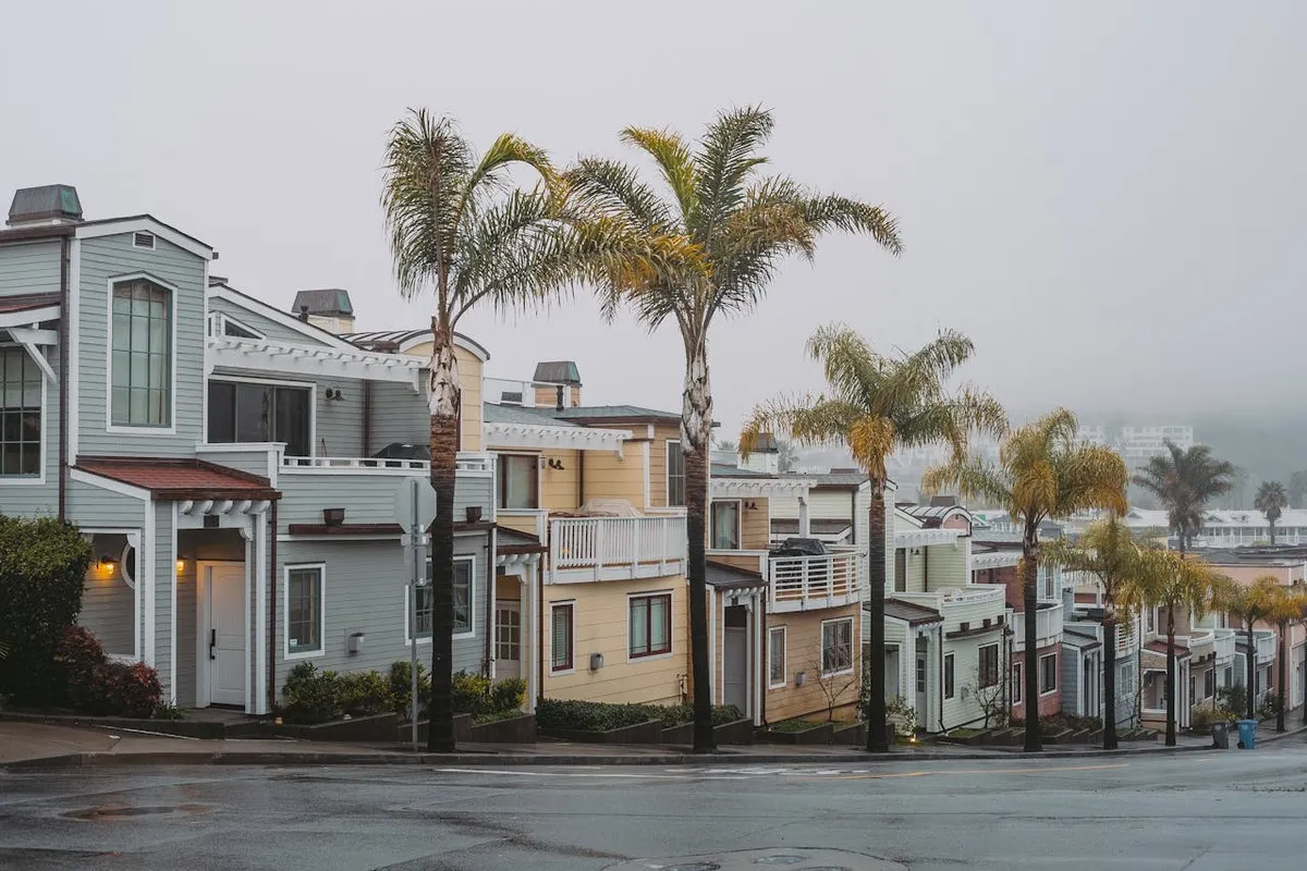 Fileira de casas geminadas (townhouses) em cores pastel na encosta, com varandas e telhados inclinados, separadas por palmeiras, em um dia nublado ou neblina (fog).