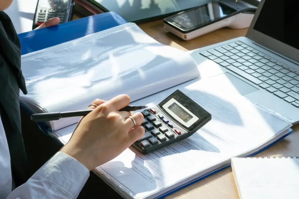 Pessoa usando uma calculadora sobre uma pilha de documentos e pastas em uma mesa de escritório, revisando finanças e contratos para a compra de um consórcio contemplado.