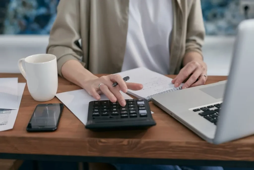 Pessoa usando uma calculadora e um laptop em uma mesa de madeira, fazendo cálculos e revisando documentos financeiros para comprar um consórcio contemplado.