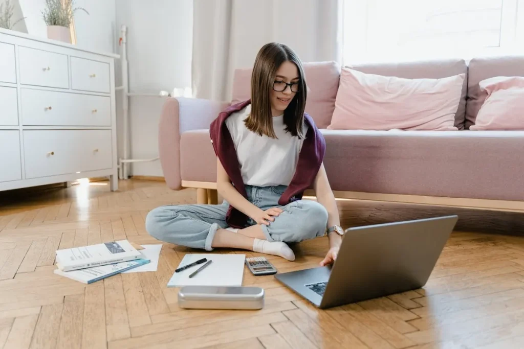 Jovem sorridente sentada no chão de madeira com laptop e calculadora, pesquisando e planejando a compra de um consórcio contemplado.