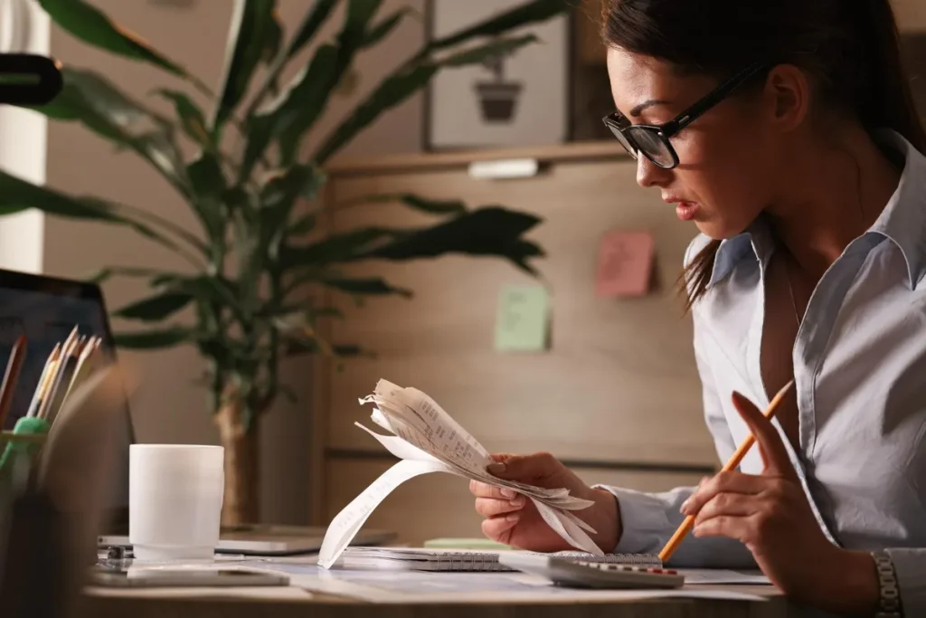 Mulher de óculos, sentada à mesa de escritório, olhando atentamente para notas e comprovantes de gastos enquanto usa um lápis e calculadora, representando controle financeiro e planejamento de consórcio.