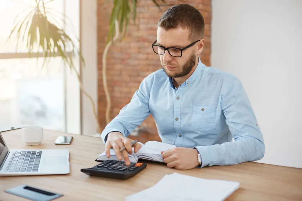 Homem branco de camisa azul e óculos, sentado à mesa com notebook e caneca de café, usando uma calculadora para anotar valores em um caderno, representando planejamento e simulação de consórcio.