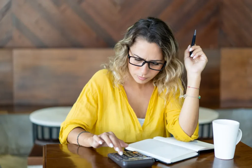 Mulher de camisa amarela e óculos, com expressão de concentração, sentada em mesa de madeira, usando uma calculadora com caneta e caderno para planejar orçamento e consórcio de 50 mil reais.