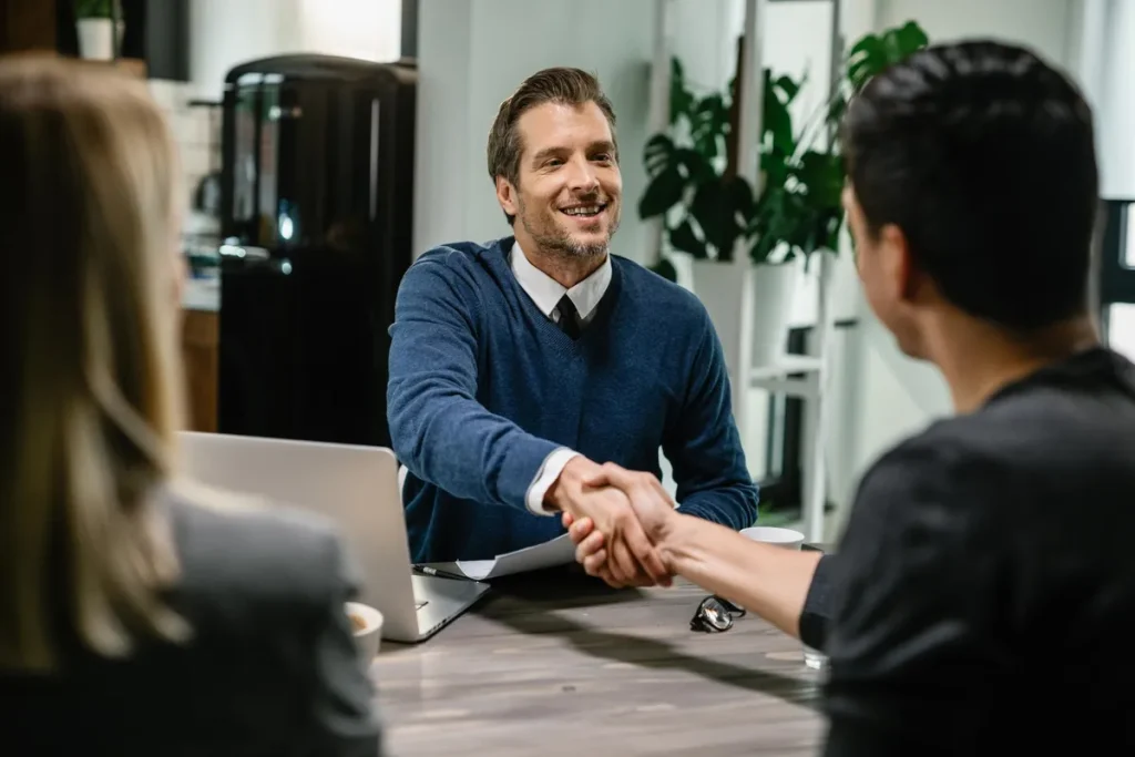 Homem sorridente em suéter azul apertando a mão de um cliente em uma mesa de reunião, com uma mulher parcialmente visível, simbolizando o início ou fechamento de um contrato de consórcio.