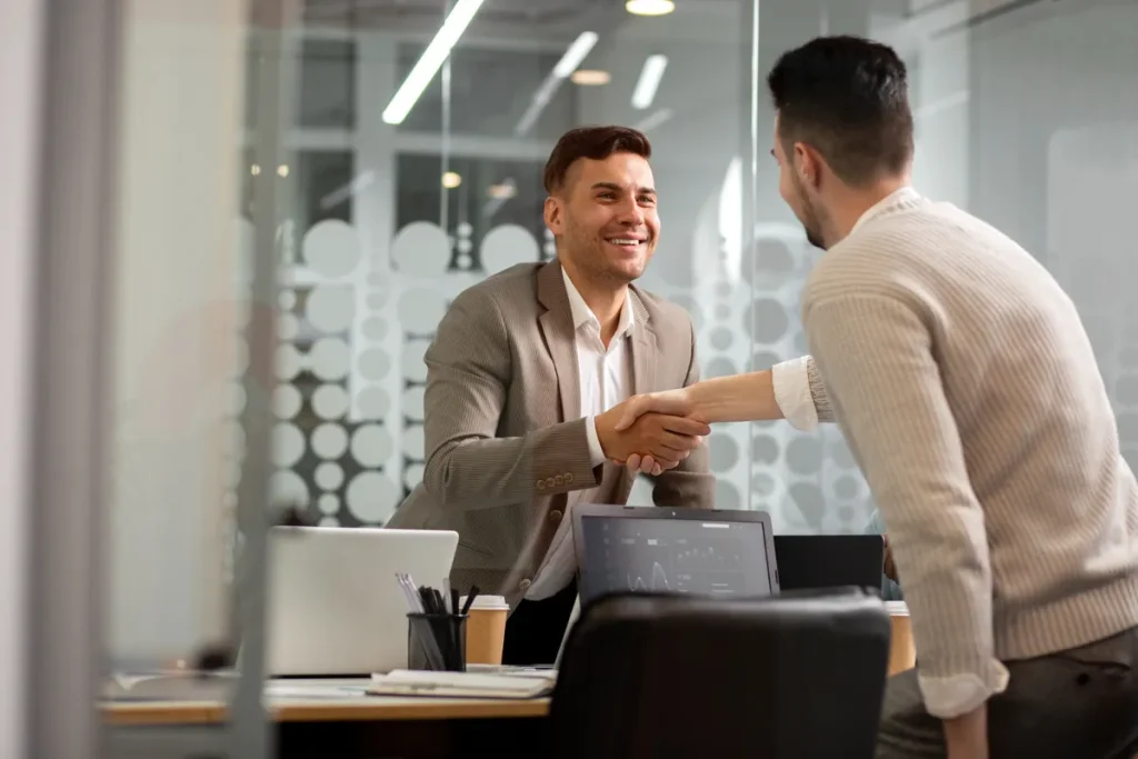 Dois homens de negócios apertando as mãos com sorrisos em um escritório moderno, simbolizando um acordo de consórcio ou parceria de negócios.