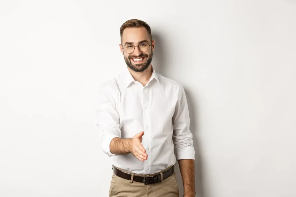 Homem profissional sorridente de óculos e camisa branca estendendo a mão para um aperto de mãos, simbolizando boas-vindas ou o início de um consórcio.