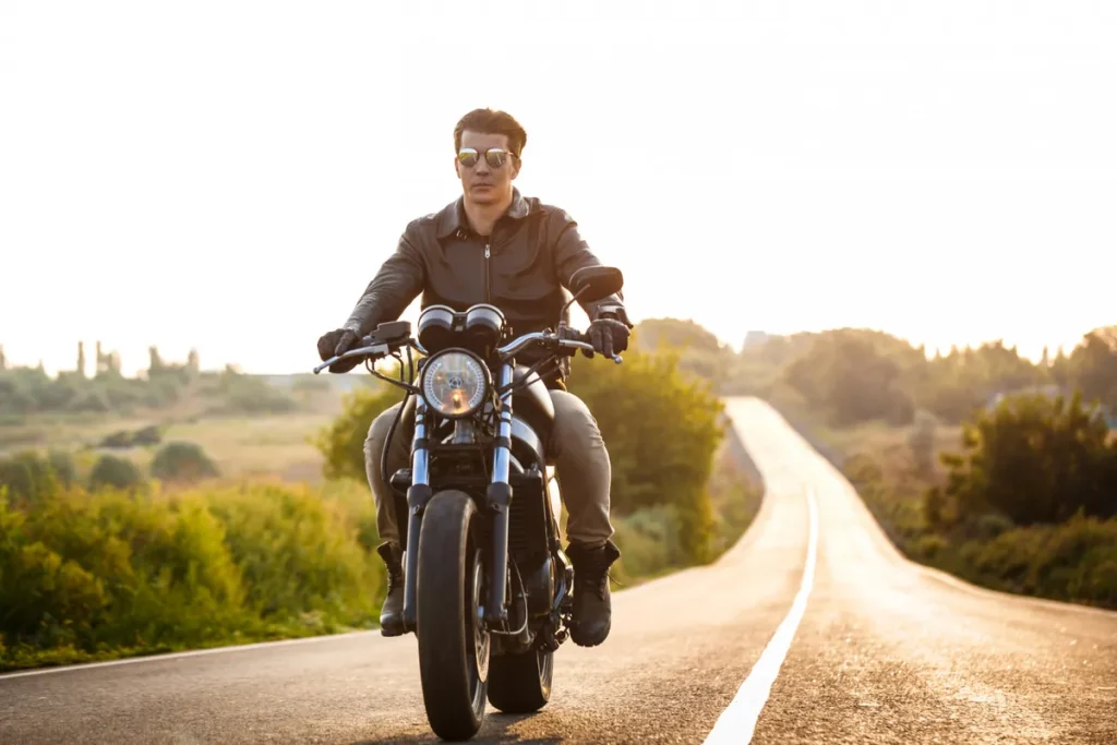 Homem de óculos de sol e jaqueta de couro pilotando uma motocicleta cruiser preta em uma estrada rural ao pôr do sol, ilustrando a conquista da liberdade com consórcio de motos.