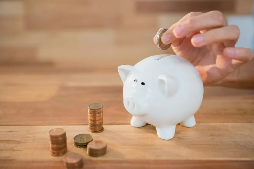 Mão colocando moeda em cofrinho branco em formato de porquinho, representando estabilidade financeira.