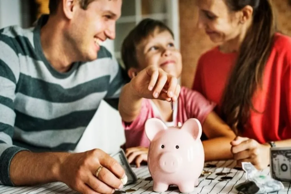 Família sorridente depositando moedas em um cofrinho rosa, simbolizando estabilidade financeira.