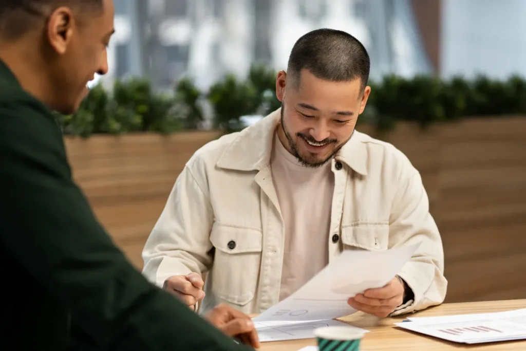Homem asiático sorridente revisando documentos com colega, discutindo finanças ou investimento.