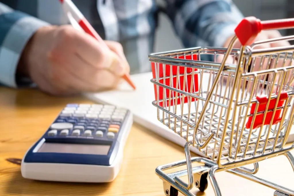 Pessoa calculando despesas com calculadora e anotações ao lado de um carrinho de compras em miniatura, simbolizando inflação e aumento do custo de vida.