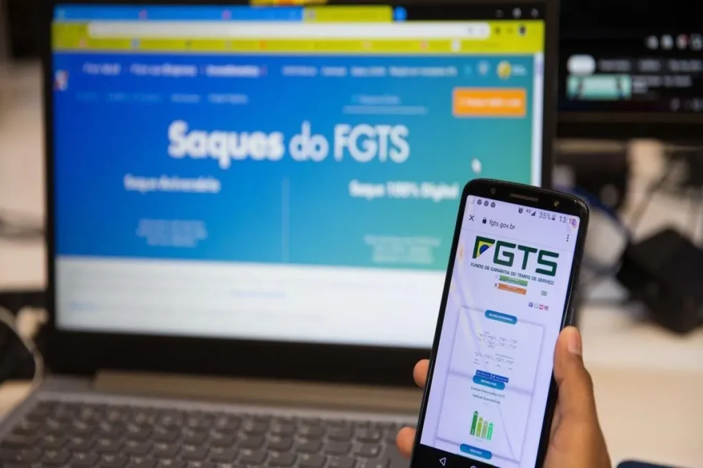 Pessoa acessando o site oficial do FGTS no celular, com computador ao fundo exibindo a página de saques do FGTS.