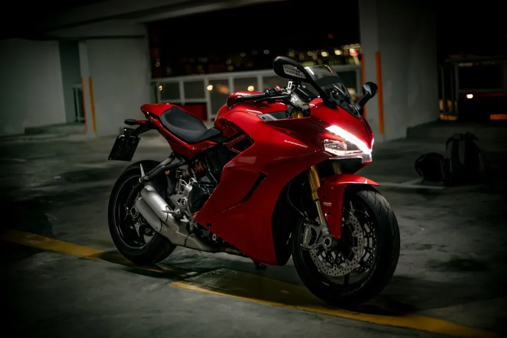 Motocicleta superesportiva vermelha (Ferrari Red) com carenagem completa, suspensão dourada e faróis de LED acesos, estacionada em estacionamento escuro.