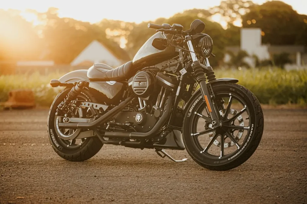Motocicleta cruiser preta (possivelmente Harley-Davidson Iron 883 ou similar) com pneus largos e design minimalista, estacionada ao pôr do sol em estrada de asfalto, representando a compra de uma moto zero.