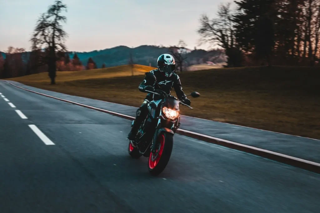 Motociclista com roupa preta pilotando uma moto naked esportiva de cor escura com detalhes vermelhos (aros da roda), em uma estrada asfaltada e sinuosa ao pôr do sol.