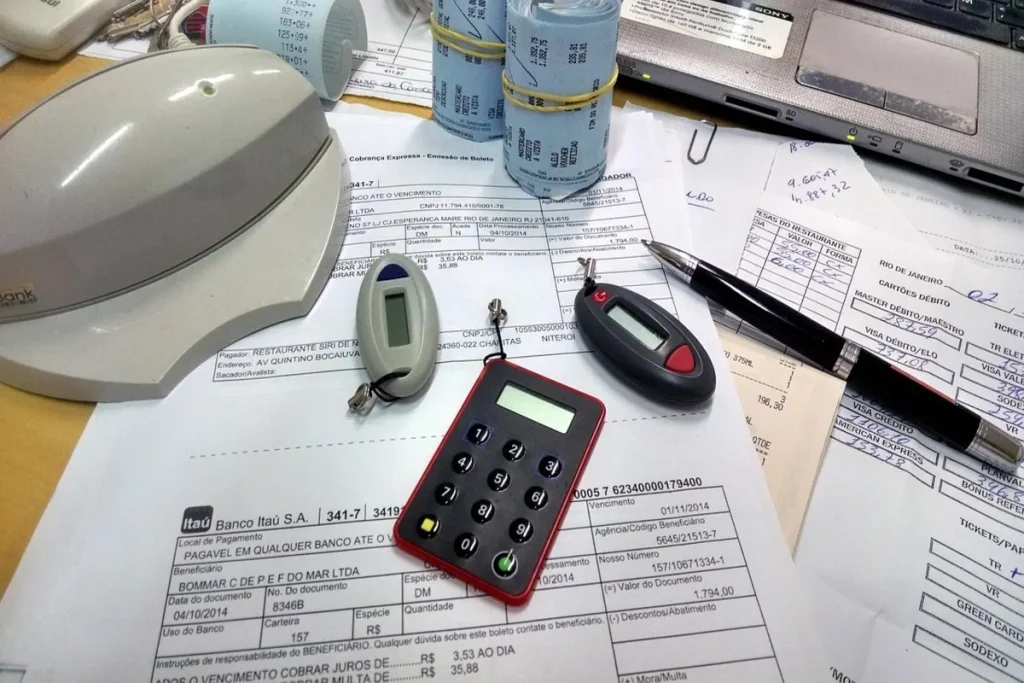 Vista de uma mesa de trabalho cheia de documentos, boletos bancários, recibos, uma calculadora de autenticação bancária e uma caneta. A imagem sugere a gestão de contas, pagamento de dívidas e rotinas de organização financeira.