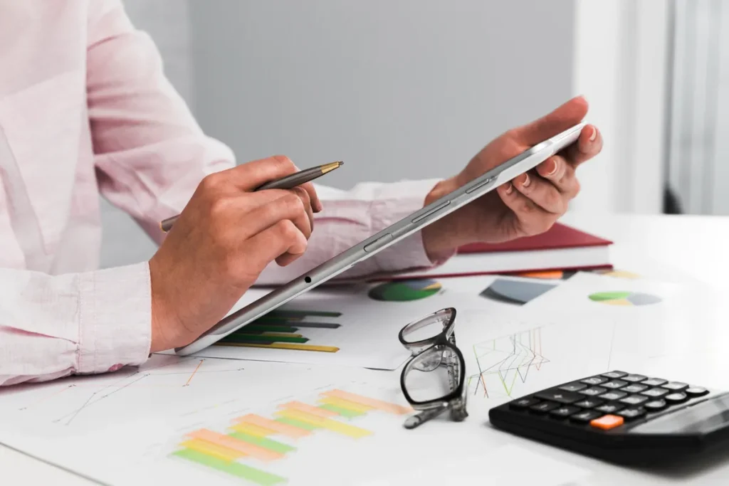 Uma pessoa de camisa rosa está segurando um tablet e uma caneta, com a mesa cheia de documentos com gráficos, uma calculadora e óculos de grau. A imagem sugere a organização de finanças, planejamento financeiro e análise de dados.