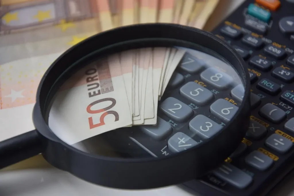 Uma lupa de aumento sobre um maço de cédulas de 50 euros. Uma calculadora preta está no plano de fundo. A imagem sugere uma análise financeira detalhada, auditoria de contas e foco em dinheiro e investimentos.