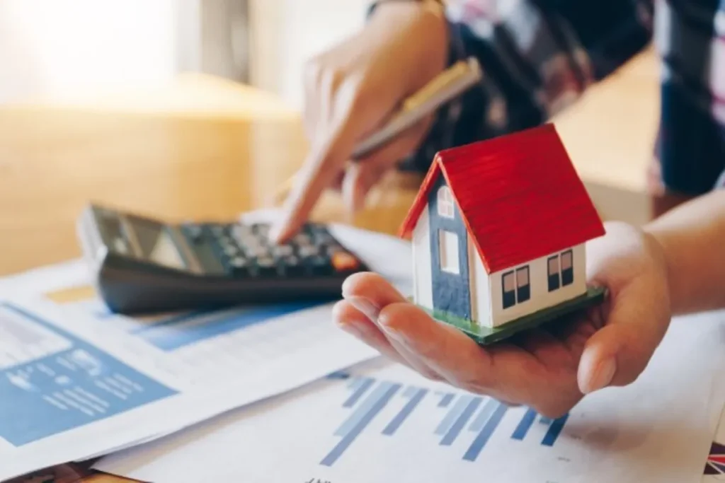 Pessoa calculando parcelas de financiamento imobiliário enquanto segura uma miniatura de casa.