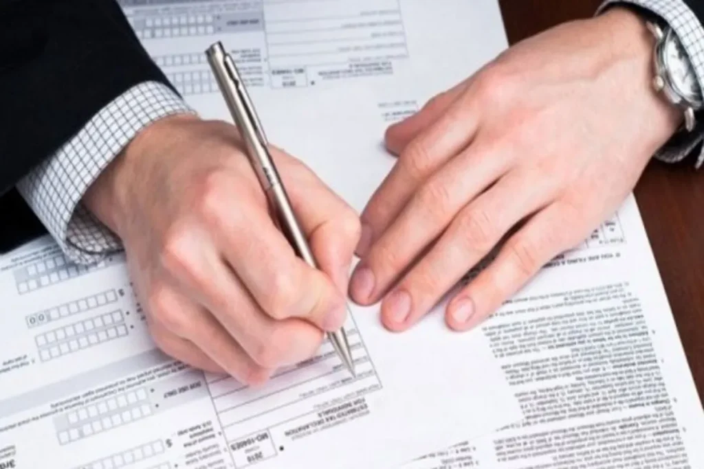 Mãos de um profissional, vestindo terno e relógio, preenchendo ou assinando formulários, representando a etapa de solicitação da certidão negativa de imóvel rural.