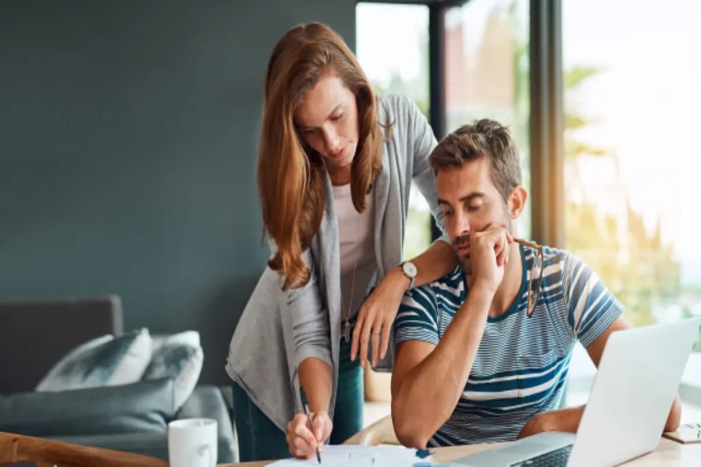 Casal jovem assinando documentos e usando um notebook em casa, simbolizando planejamento financeiro, hipoteca, ou revisão de empréstimo imobiliário.