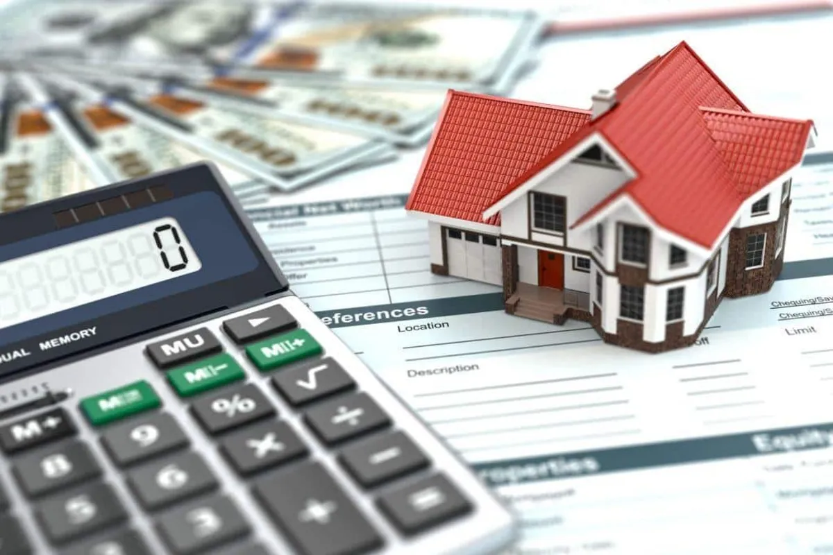 Miniatura de casa em um formulário de hipoteca/financiamento, ao lado de uma calculadora e notas de 100 dólares desfocadas, simbolizando o custo de compra de imóvel.
