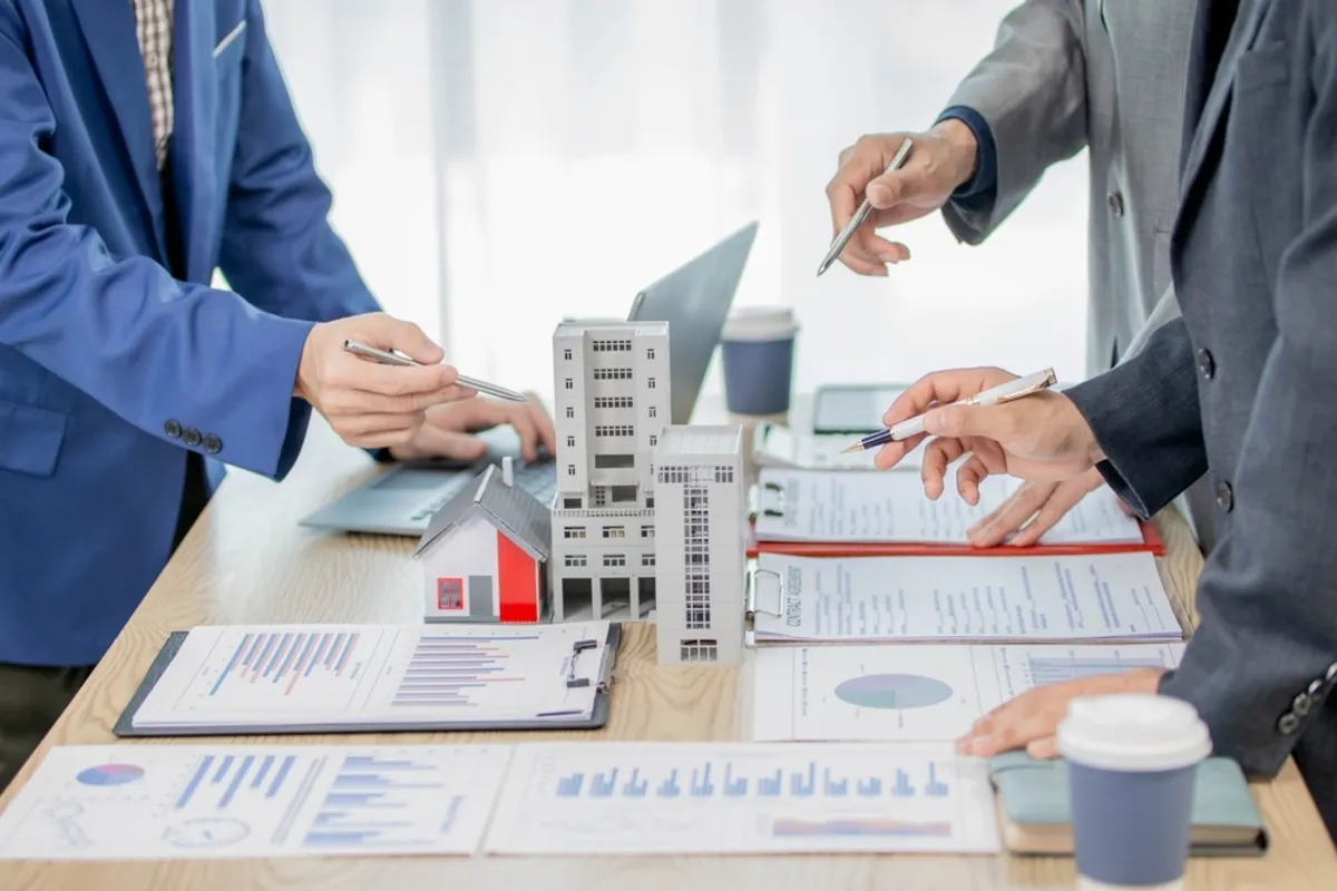 Três profissionais de terno analisando modelos de edifícios, gráficos de relatórios financeiros e contratos de incorporação imobiliária em uma mesa de reunião.
