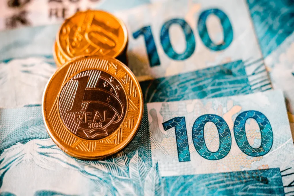 Close-up de moedas de 1 Real e notas de 100 Reais, simbolizando temas financeiros como reajuste salarial, inflação, economia e valor da moeda brasileira.