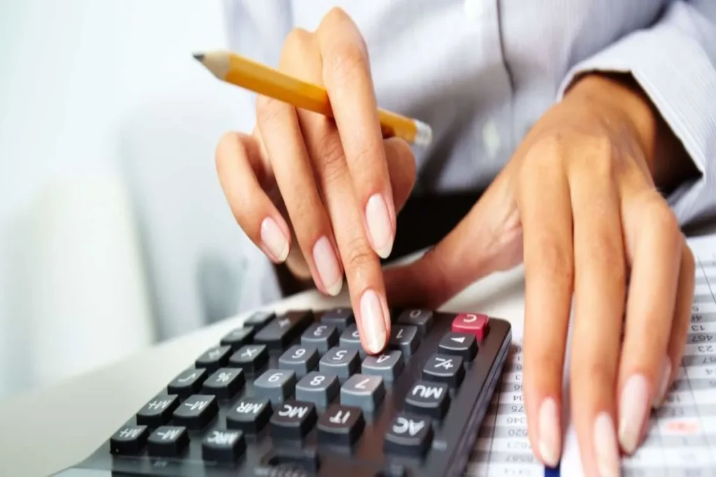 Mãos de uma pessoa usando uma calculadora com um lápis na outra mão, sobre documentos e planilhas, simbolizando cálculo, reajuste financeiro ou contabilidade.