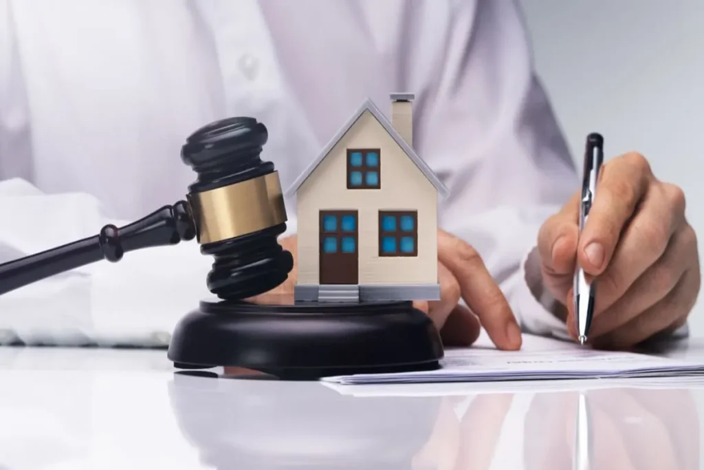 Martelo judicial (malhete) ao lado de uma maquete de casa e uma mão assinando documentos, simbolizando a segurança jurídica e a formalização da propriedade através do registro de imóvel.