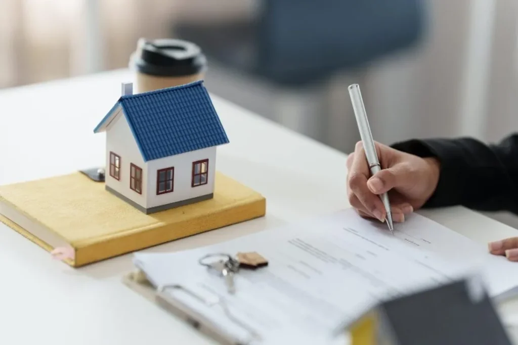 Uma pessoa assinando o documento de compra ou financiamento de uma casa, com a maquete do imóvel e as chaves à vista, representando o processo de registro de imóvel e a formalização da propriedade.