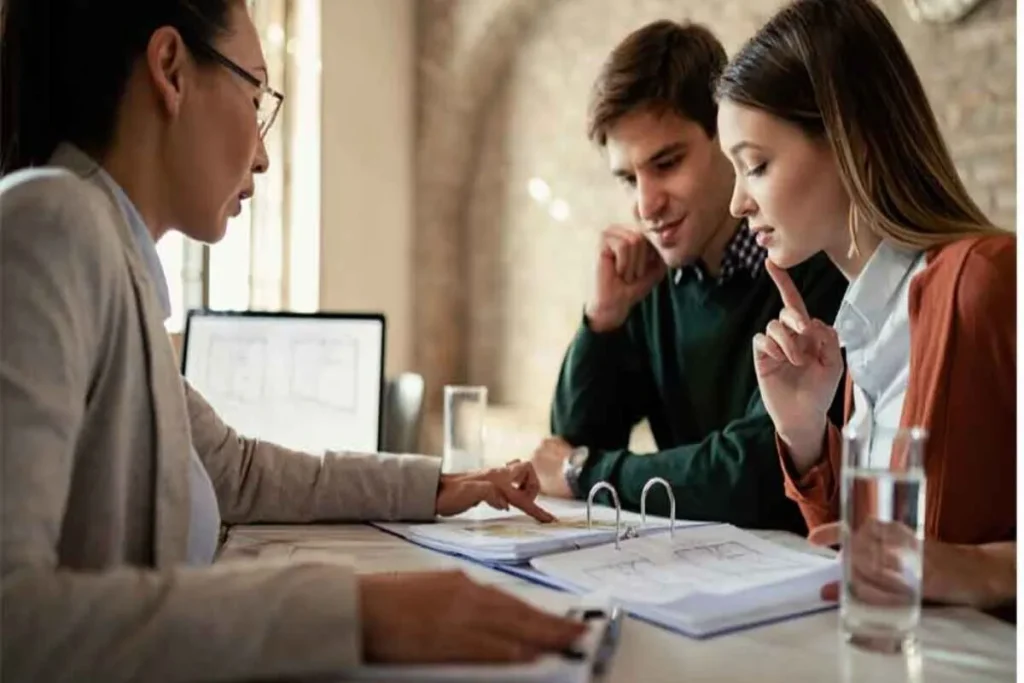 Uma consultora financeira de óculos apontando para documentos enquanto explica ao casal as cláusulas e a importância do seguro prestamista ao contratar um crédito ou financiamento.
